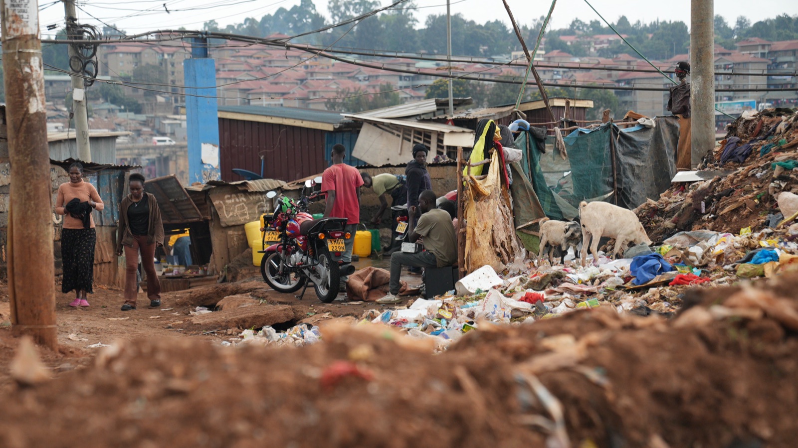 A street in Kibera at eye level: people at a motorbike and a shopfront, a heap of waste in the foreground being picked through, goats behind.
